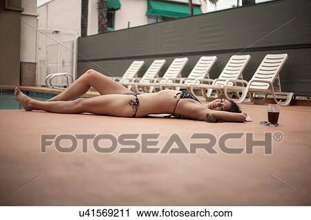 Stock Image - Young woman in bikini relaxing beside deck chairs. Fotosearch