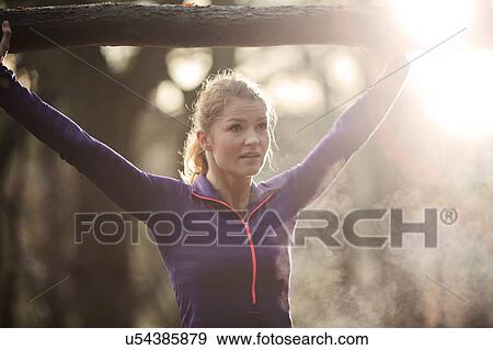 Stock Photo - Young woman in forest arms raised holding tree branch, looking away, condensation from breath. Fotosearch