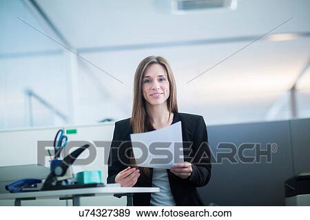 Stock Photo - Young woman in office at desk holding paperwork looking at camera smiling. Fotosearch