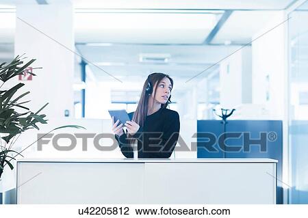 Young woman in office wearing telephone headset, holding digital tablet looking away over shoulder View Large Photo Image Stock Image - Young woman in office wearing telephone headset, holding digital tablet looking away over shoulder. Fotosearch