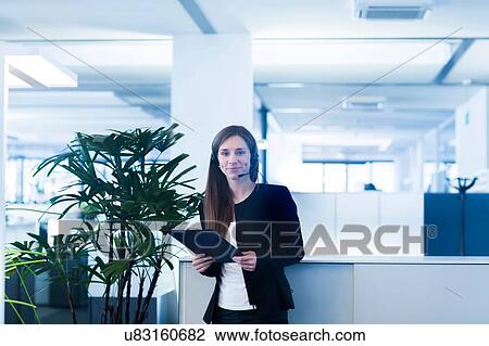 Stock Image - Young woman in office wearing telephone headset, holding digital tablet looking at camera smiling. Fotosearch