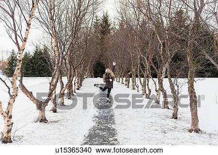Stock Image - Young woman on snow covered tree lined pathway, standing on one leg, arm raised looking up. Fotosearch