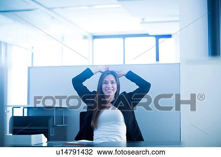 Young woman sitting at desk in office hands behind head looking at camera smiling View Large Photo Image Stock Image - Young woman sitting at desk in office hands behind head looking at camera smiling. Fotosearch