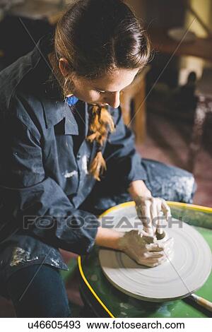 Stock Image - Young woman sitting at pottery wheel shaping clay, looking down. Fotosearch