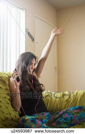 Stock Photo - Young woman sitting on sofa, wearing earbuds, listening to music, arm raised looking away smiling. Fotosearch