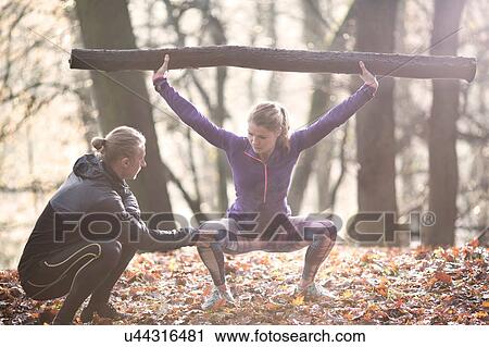 Stock Image - Young woman squatting in forest, arms raised holding tree branch. Fotosearch