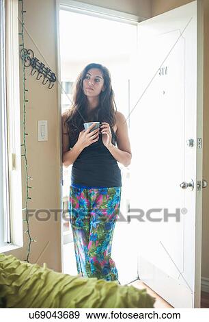 Stock Photo - Young woman standing in doorway holding coffee cup looking away. Fotosearch