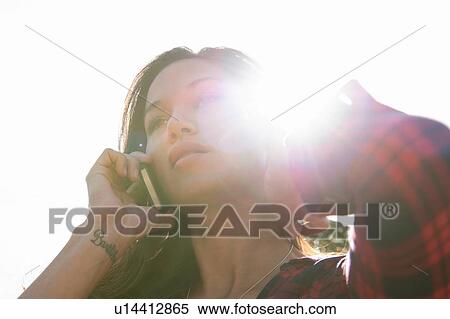 Stock Photography - Young woman talking on smartphone against sunlit sky. Fotosearch