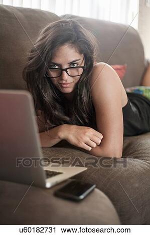Young woman wearing eyeglasses lying on front on sofa using laptop computer, looking at camera View Large Photo Image Stock Image - Young woman wearing eyeglasses lying on front on sofa using laptop computer, looking at camera. Fotosearch