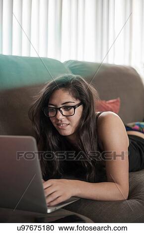 Stock Image - Young woman wearing eyeglasses lying on front on sofa using laptop computer. Fotosearch