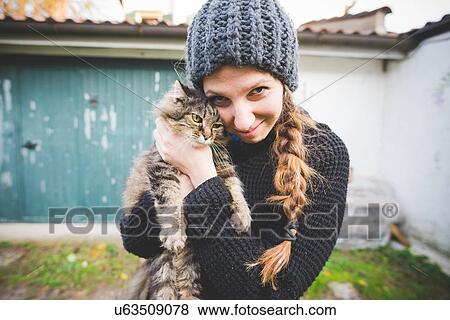 Stock Photo - Young woman wearing knit hat snuggling cat, looking at camera smiling. Fotosearch
