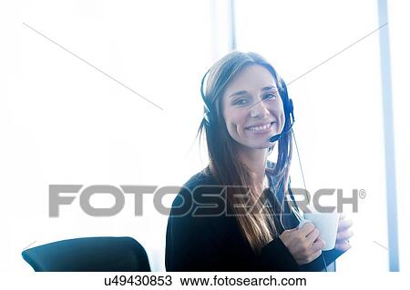 Stock Image - Young woman wearing telephone headset holding coffee cup looking at camera smiling. Fotosearch