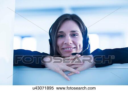 Stock Photography - Young woman wearing telephone headset looking over partition screen at camera smiling. Fotosearch