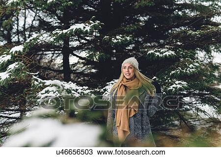 Stock Image - Young woman wearing woollen hat and scarf by snow covered tree looking away smiling. Fotosearch