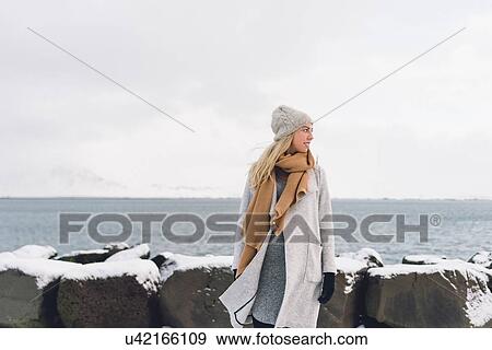 Young woman wearing woollen hat and scarf by snow covered rocks and water looking away smiling View Large Photo Image Stock Photo - Young woman wearing woollen hat and scarf by snow covered rocks and water looking away smiling. Fotosearch