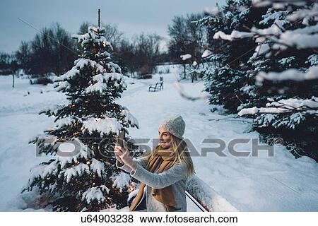 Stock Photo - Young woman wearing woollen hat and scarf by illuminated christmas tree using smartphone to take selfie smiling. Fotosearch
