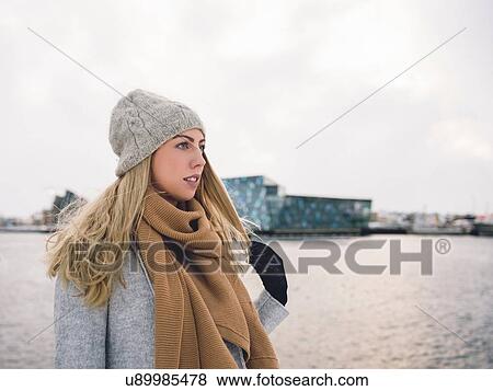 Young woman wearing woollen hat and scarf by water holding hair looking away View Large Photo Image Stock Photo - Young woman wearing woollen hat and scarf by water holding hair looking away. Fotosearch