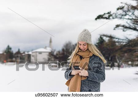 Stock Photo - Young woman wearing woollen hat and scarf on snow covered landscape holding smartphone looking down smiling. Fotosearch