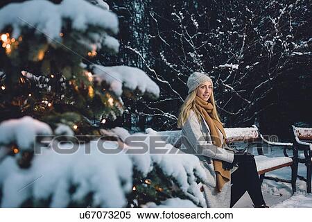 Stock Photography - Young woman wearing woollen hat and scarf sitting on snow covered park bench by illuminated christmas tree looking away smiling. Fotosearch