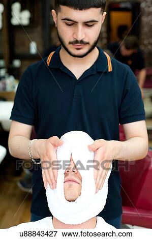 Stock Photo - Barber wrapping young mans face in fresh towel, looking down. Fotosearch