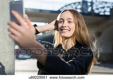 Stock Image - Beautiful mid adult woman taking smartphone selfie on city bridge. Fotosearch