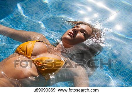 Stock Photo - Beautiful young woman floating in sunlit swimming pool wearing yellow bikini. Fotosearch
