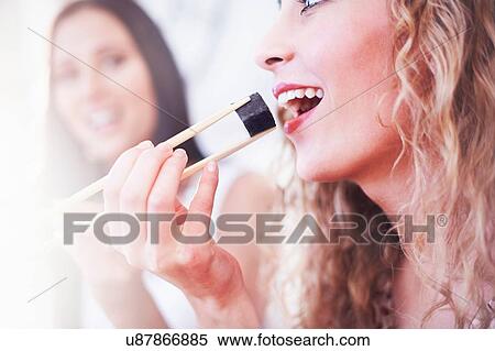 Stock Photography - Close up of two female friends eating sushi with chopsticks in kitchen. Fotosearch