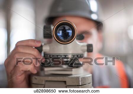 Stock Image - Close up of young male surveyor looking through theodolite on construction site. Fotosearch