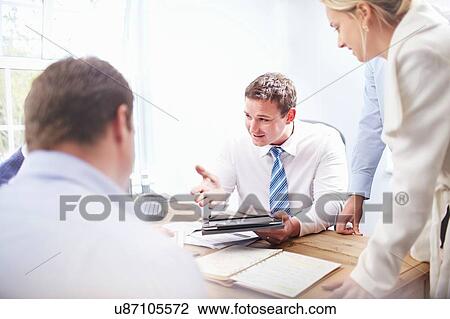 Stock Image - Colleagues in office at desk holding digital tablet having discussion. Fotosearch