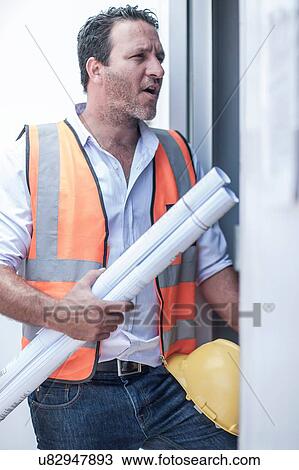 Stock Image - Construction foreman with blueprints at doorway of portable cabin on construction site. Fotosearch