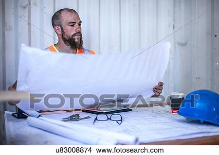 Picture - Construction worker sitting at desk looking at blueprint in portable cabin. Fotosearch