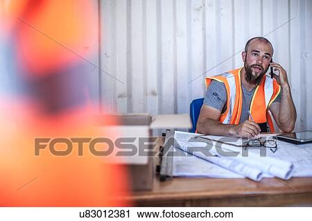 Construction worker sitting at desk talking on smartphone in portable cabin View Large Photo Image Stock Image - Construction worker sitting at desk talking on smartphone in portable cabin. Fotosearch