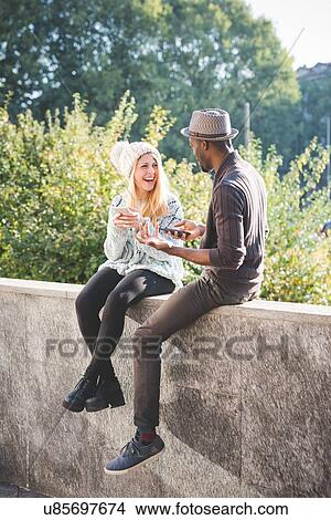 Picture - Couple chatting and using smartphone on top of cement wall. Fotosearch