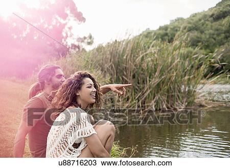 Stock Image - Couple sitting by river looking away smiling, pointing. Fotosearch