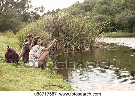 Stock Photo - Couple sitting by river looking away smiling,. Fotosearch