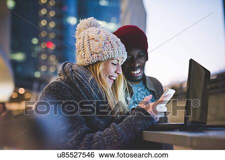 Stock Photograph - Couple using smartphone and laptop outdoors at dusk. Fotosearch