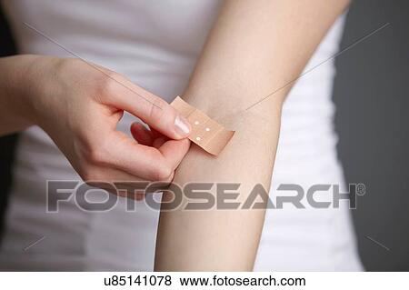 Stock Photo - Cropped close up shot of young woman applying adhesive plaster to her own arm. Fotosearch