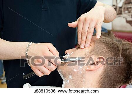 Stock Photograph - Cropped view of young man in barbershop shaving customer with straight razor. Fotosearch