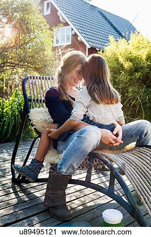 Stock Image - Daughter sitting on mother's lap on lounge chair on wooden decking. Fotosearch