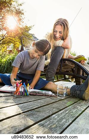 Stock Image - Daughter sitting on wooden decking drawing with mother. Fotosearch