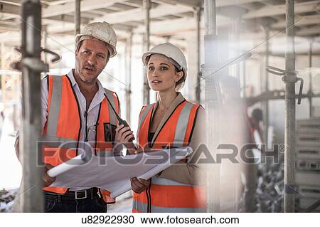 Stock Image - Female and male builders with blueprint on construction site. Fotosearch