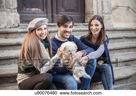 Stock Image - Friends sitting side by side on steps holding fluffy dog looking at camera smiling. Fotosearch
