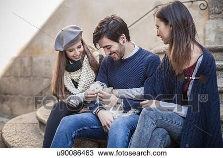Stock Image - Friends sitting side by side on steps holding fluffy dog looking down smiling. Fotosearch