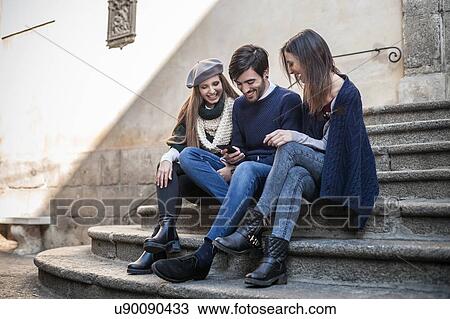 Friends sitting side by side on steps looking down at smartphone smiling View Large Photo Image Stock Image - Friends sitting side by side on steps looking down at smartphone smiling. Fotosearch