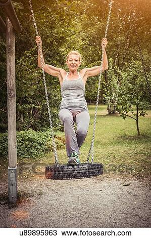 Stock Image - Full length front view of mature woman on tyre swing looking at camera smiling. Fotosearch