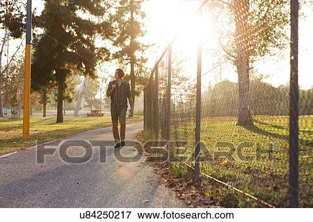 Stock Photo - Full length front view of young man walking on pathway through park looking away. Fotosearch