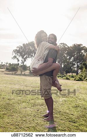 Stock Image - Full length side view of young man carrying young woman looking at camera smiling. Fotosearch