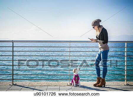 Stock Photograph - Full length side view of young woman with dog by railings in front of ocean using smartphone. Fotosearch