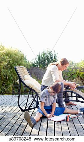 Stock Photo - Girl drawing on decking with mother sitting on lounge chair. Fotosearch