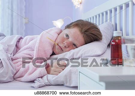 Stock Image - Girl lying in bed with thermometer in her mouth. Fotosearch